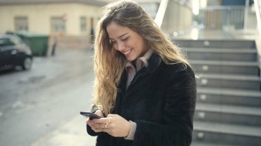 Genç, güzel, İspanyol bir kadın sokakta akıllı telefon kullanarak gülümsüyor.