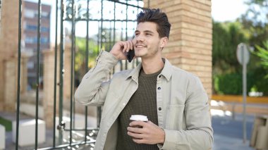 Young hispanic man talking on smartphone drinking coffee at street