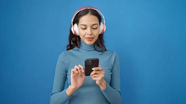 孤立した青の背景にヘッドフォンを身に着けているスマートフォンを使用して若い美しいヒスパニック女性 — ストック写真