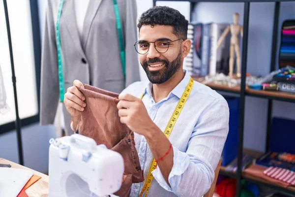 年轻的阿拉伯男子裁缝笑着自信地在裁缝店里拿着牛仔裤 — 图库照片
