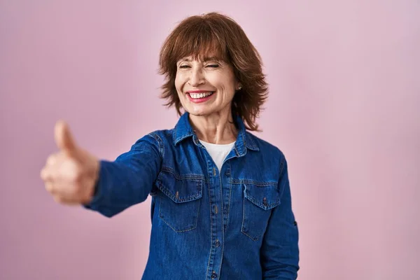 Middle age woman standing over pink background looking proud, smiling doing thumbs up gesture to the side