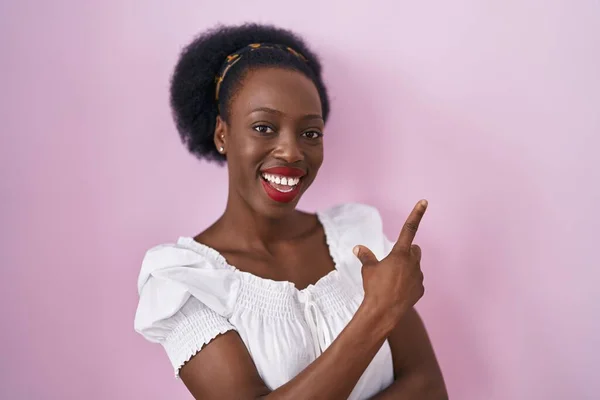 头发卷曲的非洲妇女站在粉色的背景上 脸上挂着大大的笑容 手指头指向侧面看相机 — 图库照片