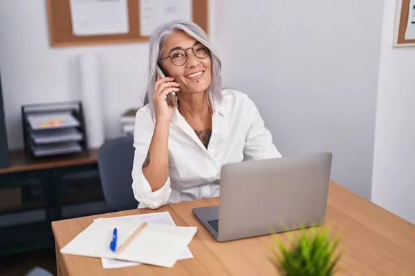 在办公室使用笔记本电脑用智能手机聊天的中年灰发女商人 — 图库照片