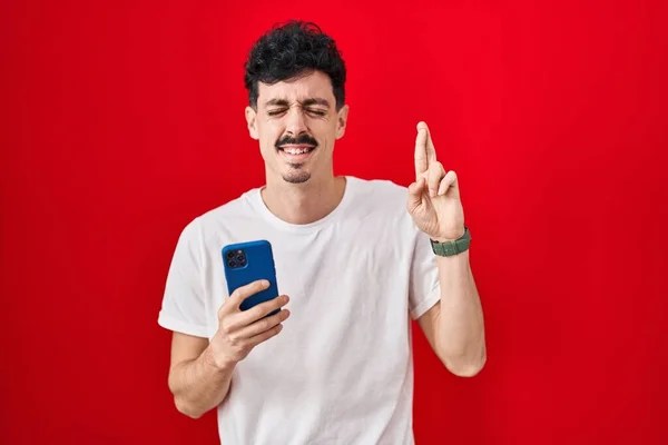 Hispanic Man Using Smartphone Red Background Gesturing Finger Crossed Smiling — Stock Photo, Image