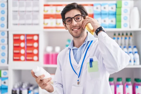 年轻的恐慌男子药剂师拿着药瓶在药店用智能手机说话 — 图库照片