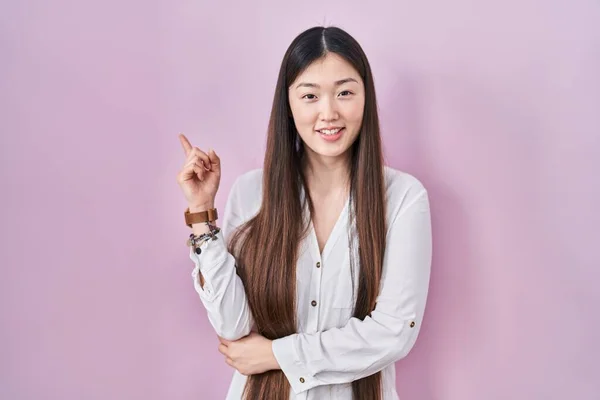 中国的年轻女子站在粉色的背景上 脸上挂着大大的笑容 手指头指向旁边看相机 — 图库照片