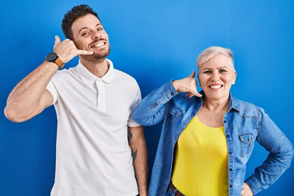 若いブラジル人の母親と息子が青い背景の上に立って 電話で話すような手と指で電話のジェスチャーをして笑っている コミュニケーションの概念 — ストック写真