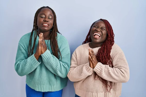 青い背景の上に立つ2人のアフリカ人女性は 非常に感情的で心配している顔に希望の表情と一緒に手で物乞いと祈りをしています お願い — ストック写真