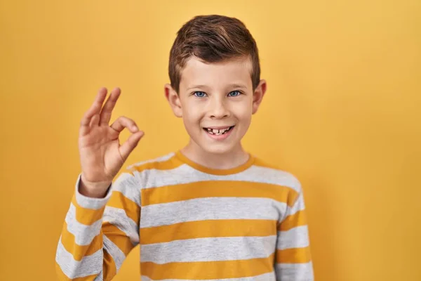 黄色の背景に立っている若い白人の子供は手と指でOkサインをして積極的に笑っています 成功した表現 — ストック写真