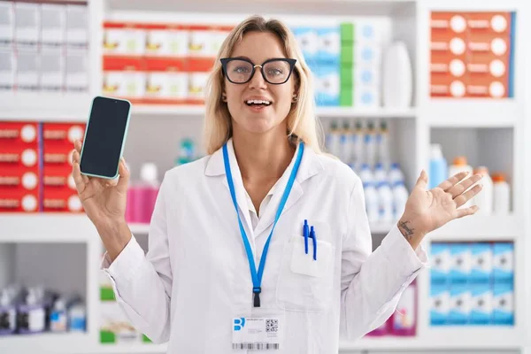 幸せな笑顔で達成を祝うスマートフォンの画面を示す薬局で働く若いブロンドの女性と手を上げて受賞式 — ストック写真