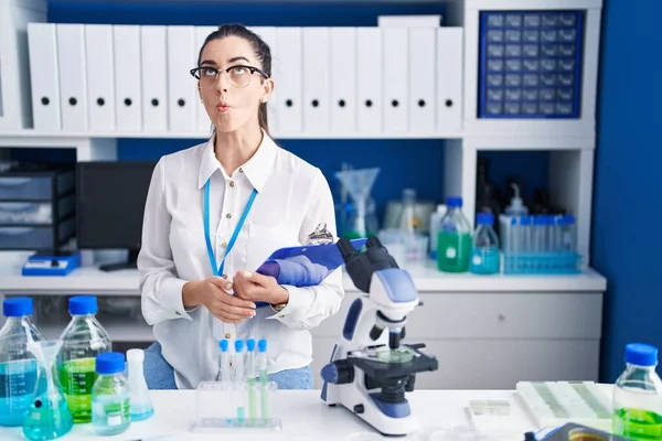 Young brunette woman working at scientist laboratory making fish face with lips, crazy and comical gesture. funny expression.