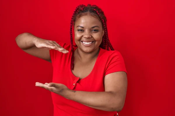 大きなと大規模なサインを示す手で赤い背景ジェスチャーの上に立って編組髪を持つアフリカ系アメリカ人女性は シンボルを測定します カメラを見て笑ってる 概念を測定し — ストック写真