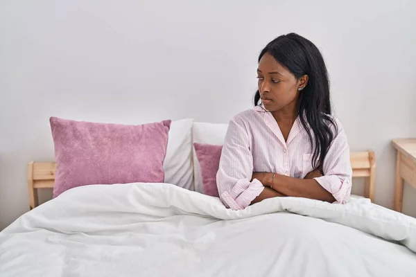 年轻的非洲裔美国女人坐在床上 脸上带着严肃的表情和双臂 在卧室里交叉着手势 — 图库照片