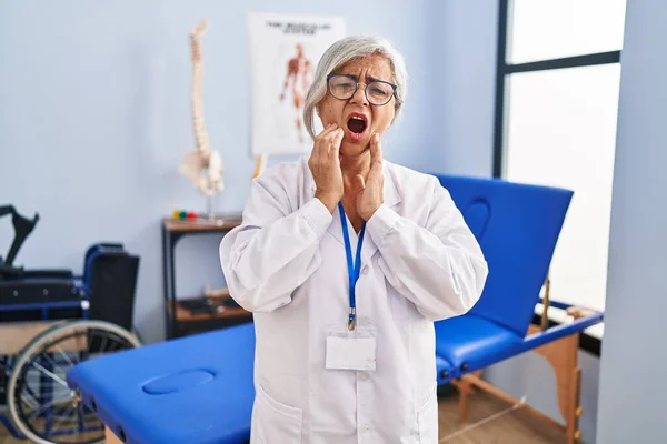 Middle age woman with grey hair working at pain recovery clinic touching mouth with hand with painful expression because of toothache or dental illness on teeth. dentist