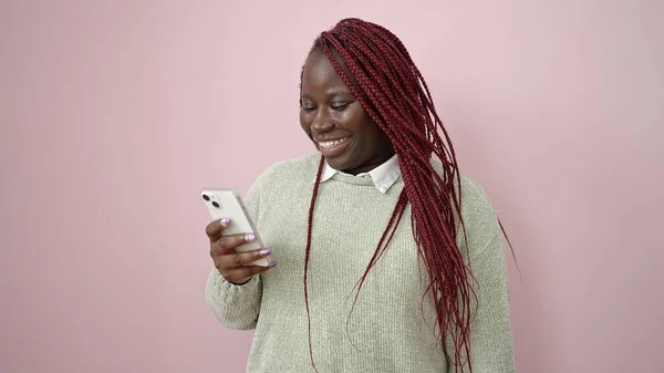在孤立的粉色背景下使用智能手机编织头发的非洲妇女 — 图库照片