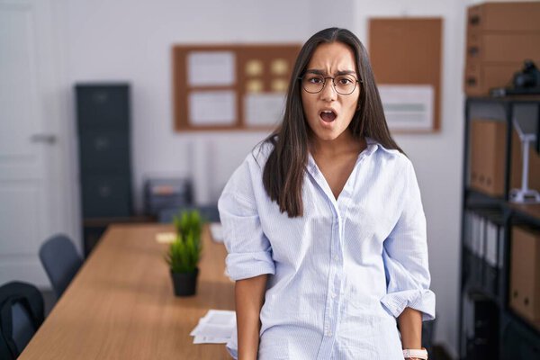 Young hispanic woman at the office in shock face, looking skeptical and sarcastic, surprised with open mouth 