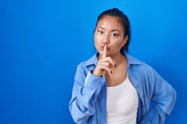 站在蓝色背景上的亚洲年轻女子要求保持安静 手指放在嘴唇上 沉默和秘密概念 — 图库照片