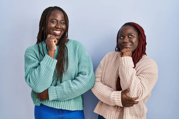 2人のアフリカ人女性が青い背景の上に立っていて 顎は質問 集中的な表現について考えている 笑顔で思いやりのある顔 疑わしい概念 — ストック写真