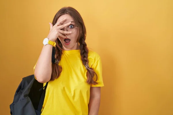 Młoda Kaukaska Kobieta Nosząca Studencki Plecak Żółtym Tle Podglądająca Dłońmi — Zdjęcie stockowe