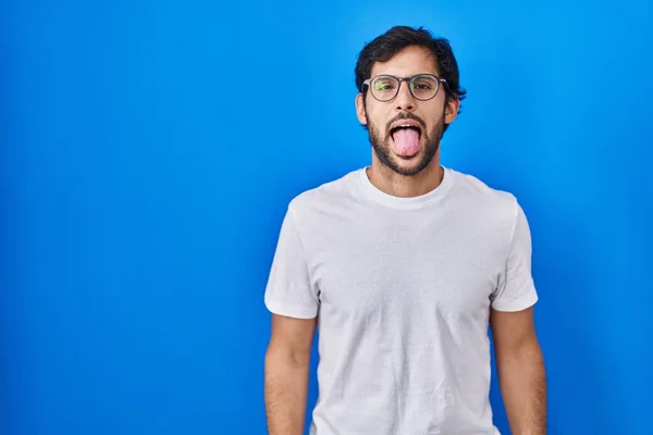 面白い表情で楽しそうに舌を突き出している青い背景の上に立つハンサムなラテン人 感情の概念 — ストック写真