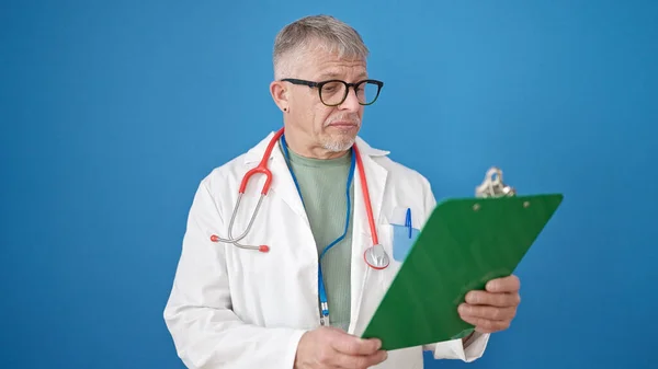中年白发苍苍的男医生站在那里 带着轻松的表情阅读着孤立的蓝色背景的文件 — 图库照片