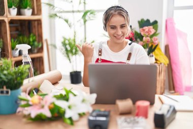Young blonde woman working at florist shop doing video call pointing thumb up to the side smiling happy with open mouth 