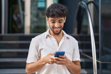 Genç Arap adam sokakta akıllı telefon kullanarak kendinden emin gülümsüyor.