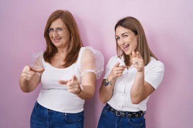 İspanyol anne ve kızı pembe arka plan üzerine gündelik beyaz tişört giyip mutlu ve komik yüzlü parmakları kameraya doğrultuyorlar. iyi enerji ve titreşimler. 
