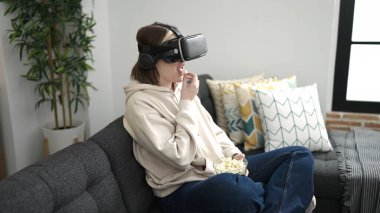 Young blonde woman watching movie using virtual reality glasses eating popcorn at home