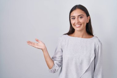 Young hispanic woman standing over white background smiling cheerful presenting and pointing with palm of hand looking at the camera. 