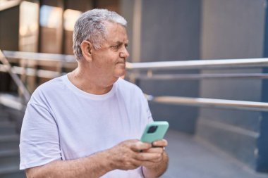 Orta yaşlı, kır saçlı bir adam sokakta rahat bir ifadeyle akıllı telefon kullanıyor.