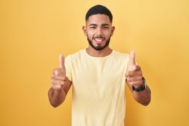 Young hispanic man standing over yellow background pointing fingers to camera with happy and funny face. good energy and vibes. 