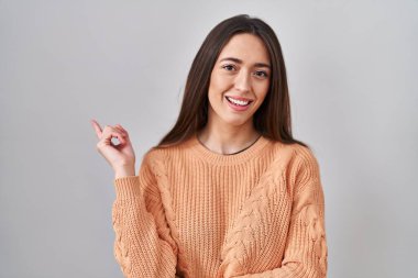 Beyaz arka planda duran genç esmer kadın, yüzünde büyük bir gülümseme ile, kameraya el ile işaret ediyor.. 