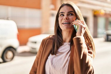 Genç, güzel, İspanyol bir kadın gülümsüyor ve caddede akıllı telefondan konuşuyor.