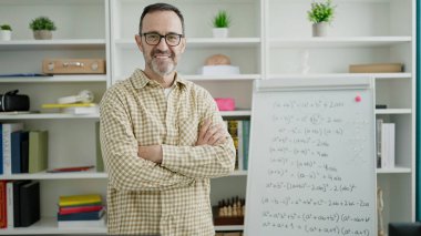 Orta yaşlı öğretmen gülümsüyor. Kollarını kavuşturup sınıfta duruyor.