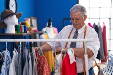 Orta yaşlı, gri saçlı, terzi dükkanında elbise askısı tutan bir adam.
