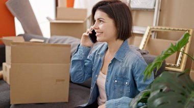 Genç beyaz kadın kanepede oturuyor ve yeni evinde telefonla konuşuyor.