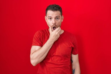 Kırmızı arka planda duran İspanyol genç adam, elleri çenesinde, inanamama, şaşkınlık ve şaşkınlık içinde bakıyor. 