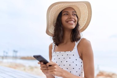 Young african american woman wearing summer hat using smartphone at seaside