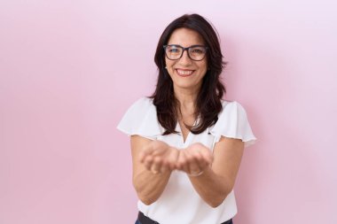 Orta yaşlı İspanyol kadın, günlük beyaz tişört ve gözlük takıyor, avuçlarıyla gülümsüyor, el hareketi yapıyor. Bekleme ve koruma 