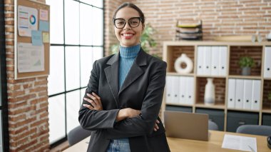 Genç ve güzel İspanyol iş kadını. Ofiste kollarını kavuşturarak gülümsüyor.
