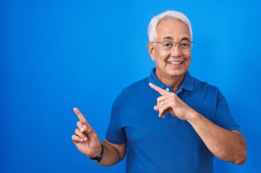 Middle age man with grey hair standing over blue background smiling and looking at the camera pointing with two hands and fingers to the side. 