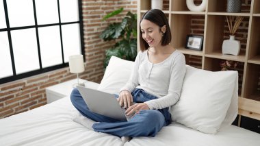 Young beautiful hispanic woman using laptop sitting on bed at bedroom