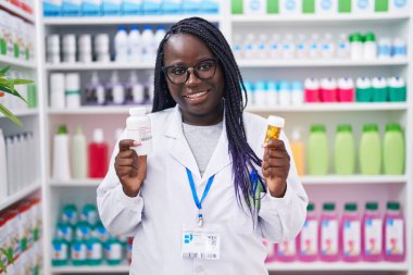 African american woman pharmacist smiling confident holding pills bottles at pharmacy