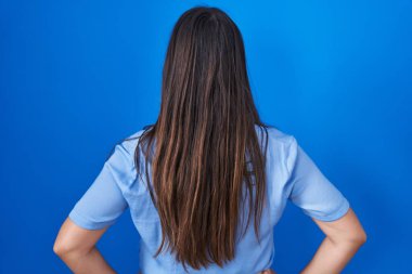 Genç esmer kadın mavi arka planda dikiliyor geriye bakıyor kolları vücudunda. 