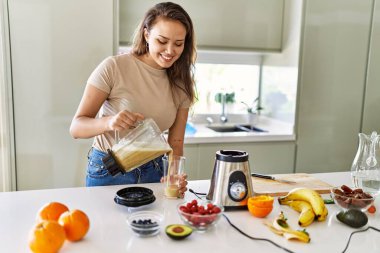 Young beautiful hispanic woman smiling confident pouring vegetable smoothie on glass at the kitchen