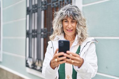 Sokakta akıllı telefon kullanan orta yaşlı bir kadın gülümsüyor.