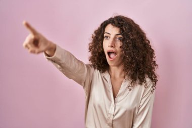 Pembe arka planda duran kıvırcık saçlı İspanyol kadın. Parmakları önde, ağzı açık, şaşırmış, ön tarafta bir şey var. 