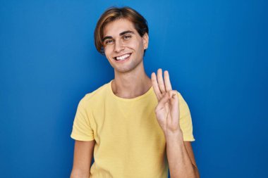 Mavi arka planda duran genç adam üç numarayı gösterip işaret ederken kendinden emin ve mutlu bir şekilde gülümsüyor.. 