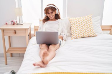 Middle age woman using laptop and headphones sitting on bed at bedroom
